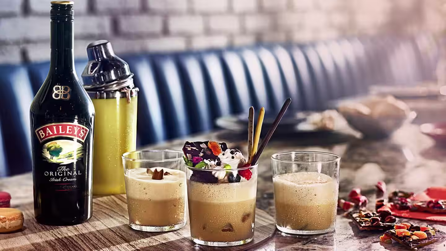 Three Baileys cocktails in glasses, one topped with whipped cream and chocolate, next to a Baileys bottle and a cocktail shaker on a bar table.