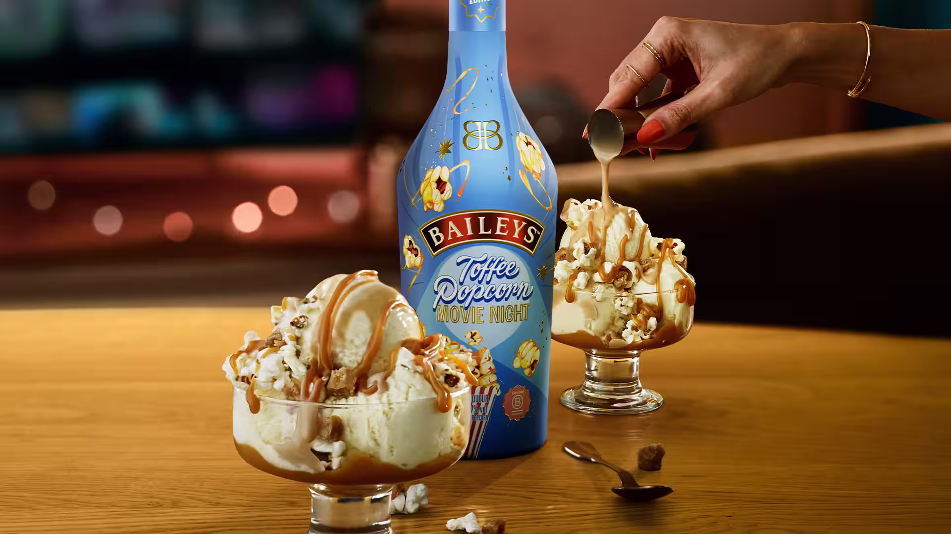 Baileys Toffee Popcorn bottle with two caramel-drizzled ice cream servings topped with whipped cream and popcorn, set for a cosy movie night scene.