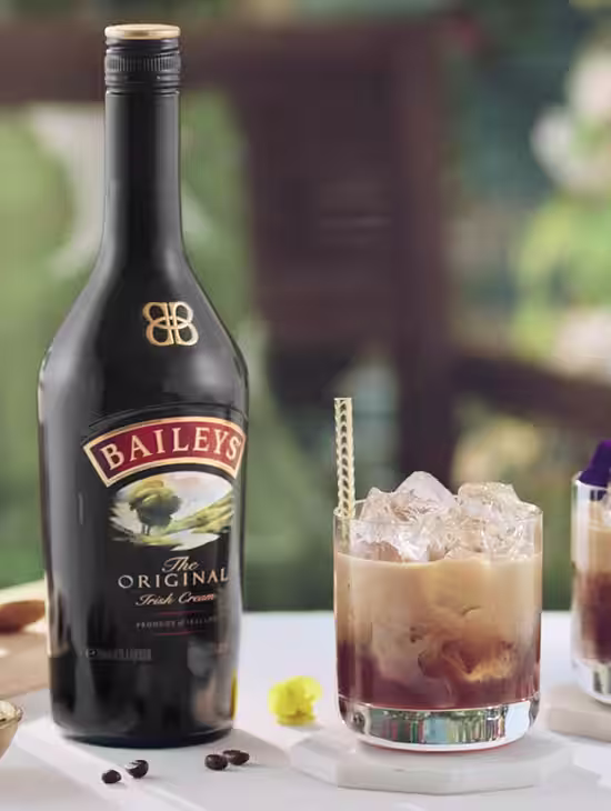A Baileys White Russian cocktail in a glass with ice, swirled with cream, next to a bottle of Baileys Irish Cream on a white table.