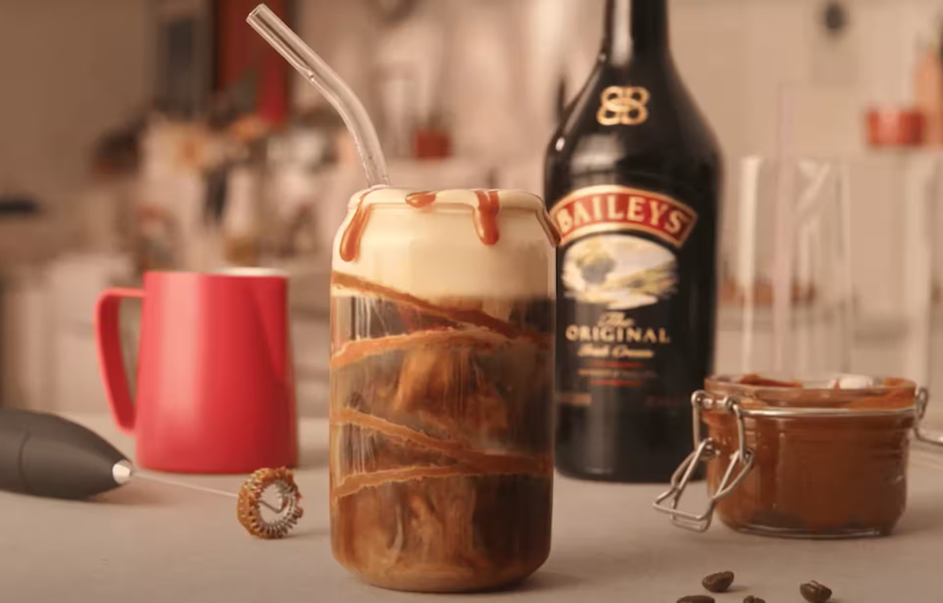 Iced coffee with Baileys cold foam in a glass, drizzled with caramel, next to a Baileys bottle, coffee beans, and a jar of caramel sauce.