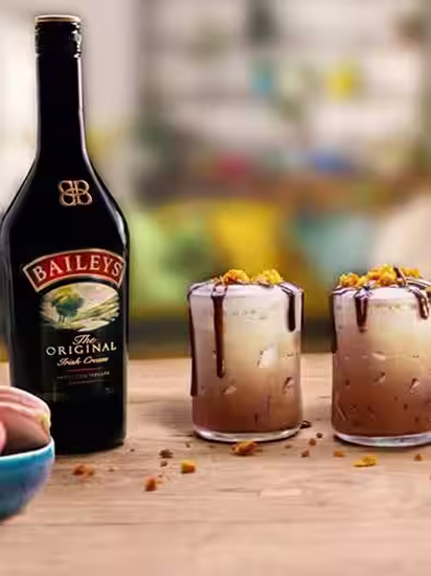 Two Baileys Chocaccino cocktails in glasses with ice, chocolate drizzle, and crumbled topping, next to a Baileys bottle on a wooden table.