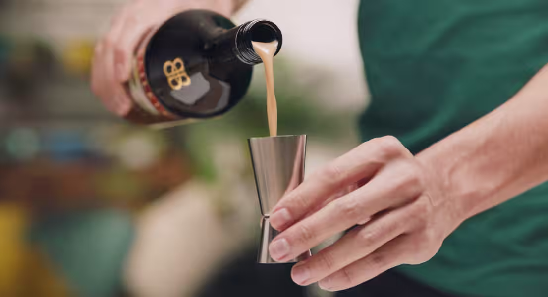 A person pours Baileys Irish Cream into a cocktail jigger.